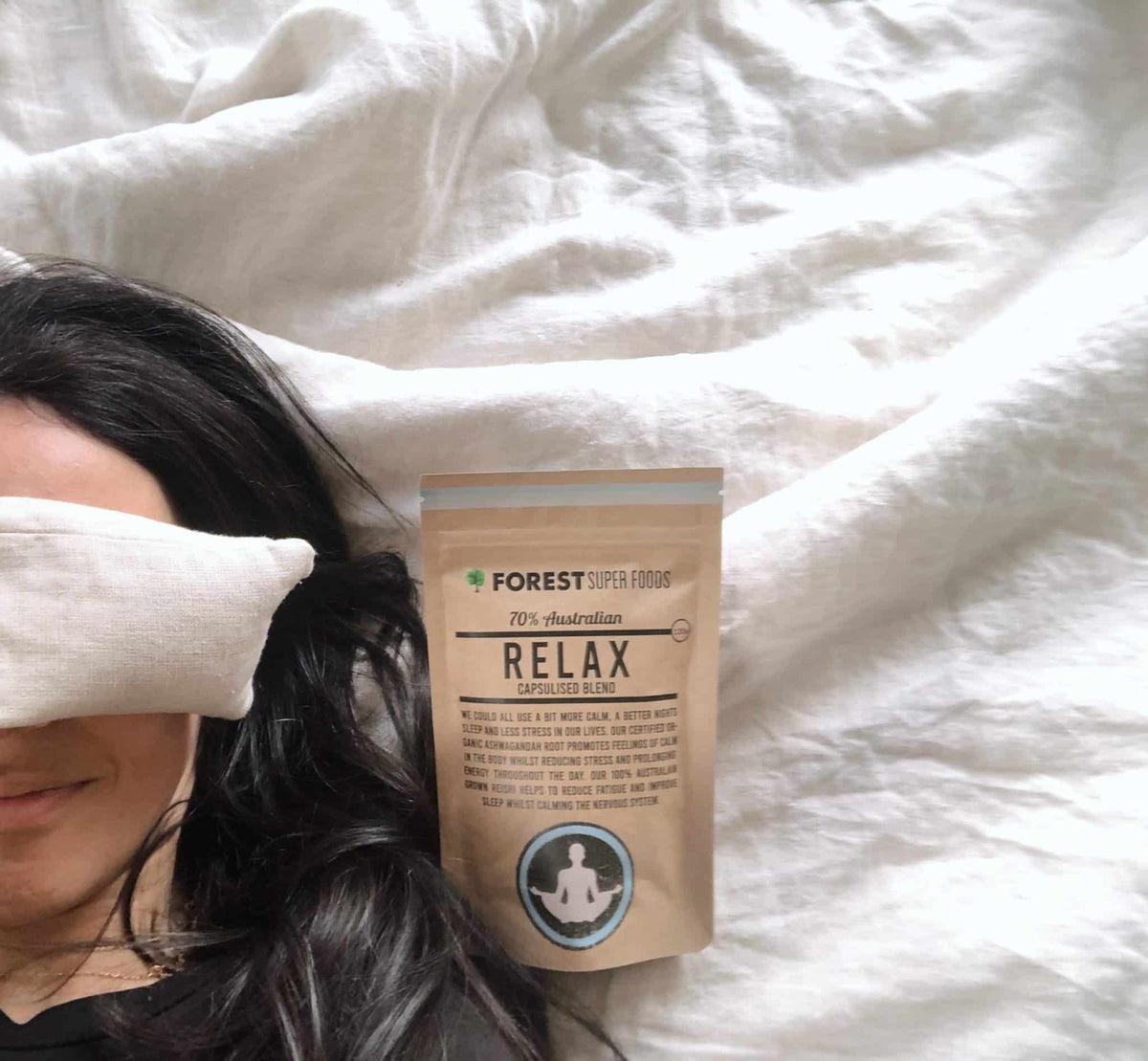 Lavender relaxation eye pillow resting on a woman’s face with a Forest Super Foods Relax herbal blends pack nearby on white bedding